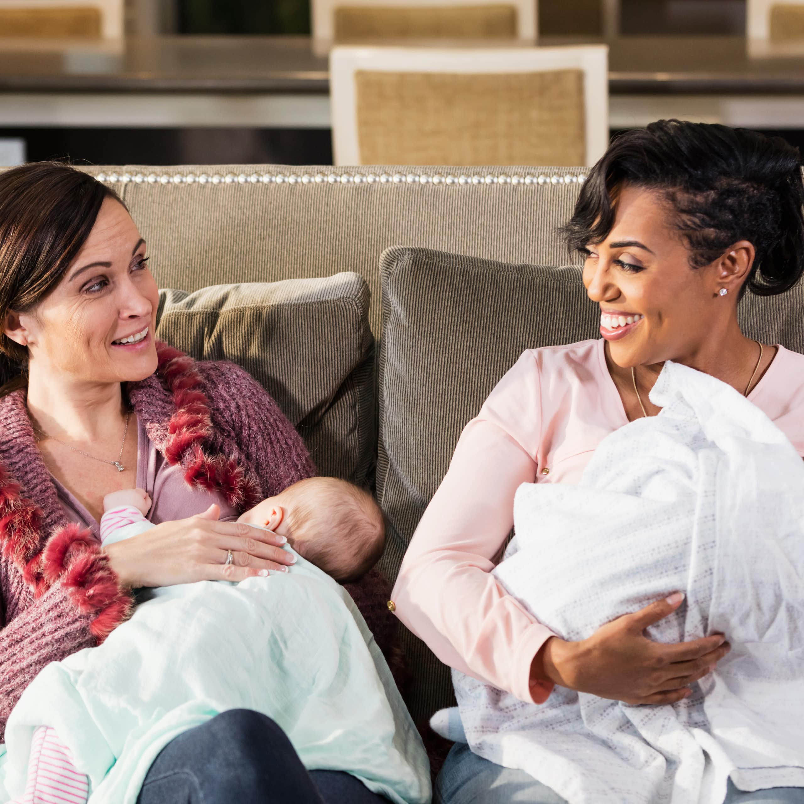Two women holding babies sit next to each other on a couch and talk.