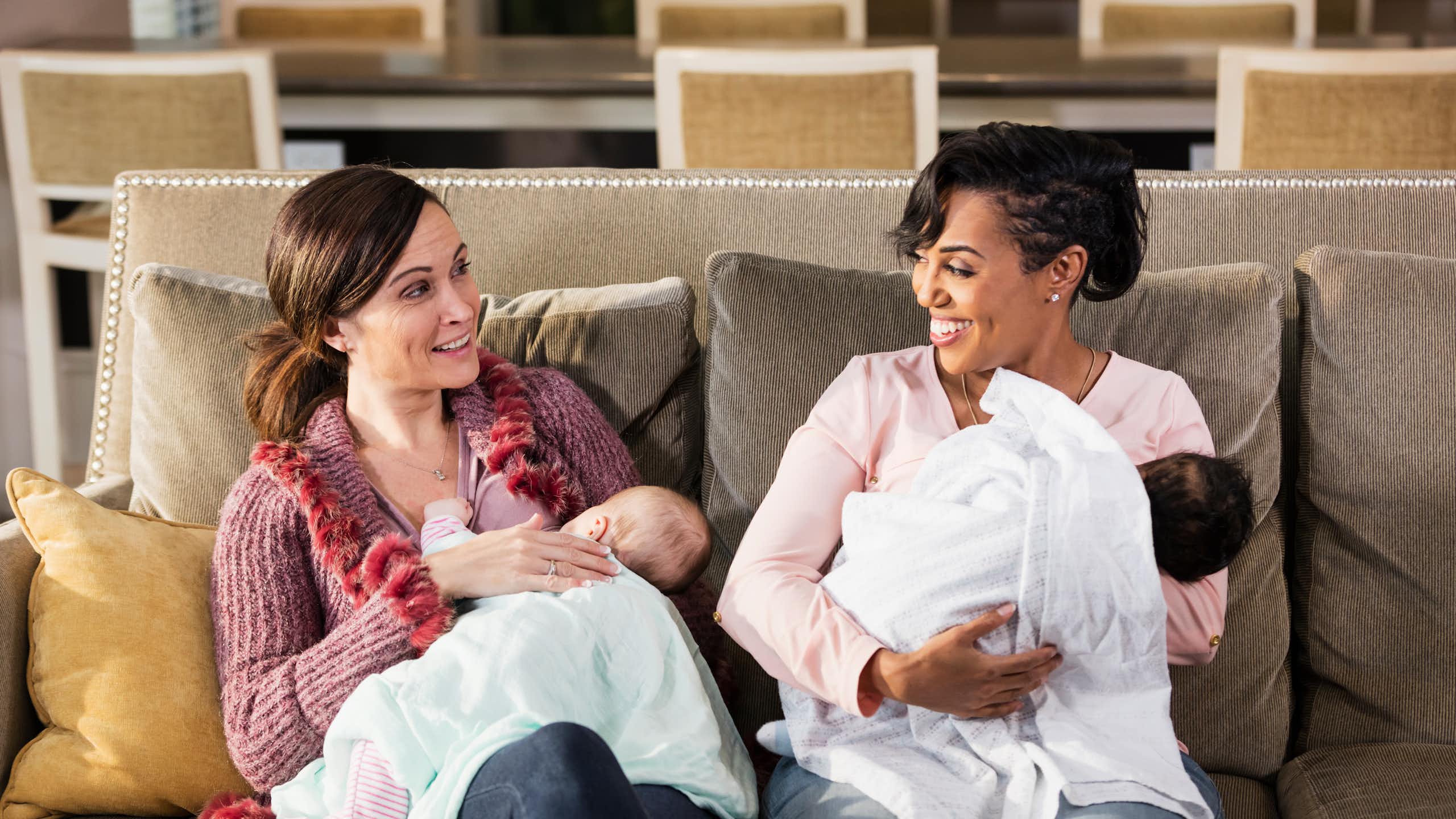 Two women holding babies sit next to each other on a couch and talk.