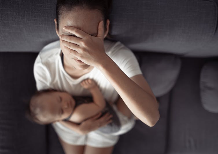 A woman holding a baby on a couch covers her eyes in distress.