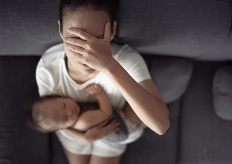 A woman holding a baby on a couch covers her eyes in distress.