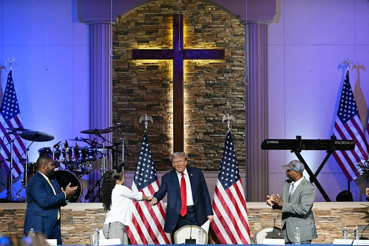 Donald Trump shakes a woman's hand in a sanctuary with a large cross and several American flags.