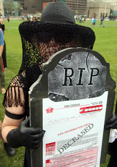 A person in a black hat holding a form that says 'deceased' and a tombstone.