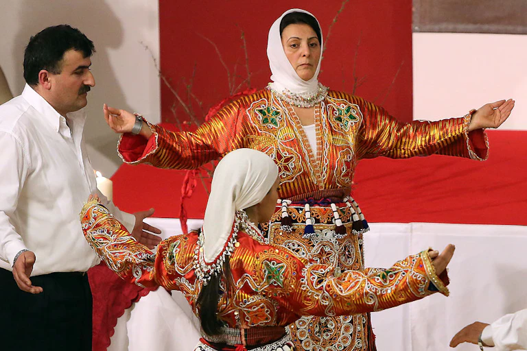 Two women in white kerchiefs and bright, ornately decorated red dresses dance with outstretched arms, with a man in a white shirt between them.
