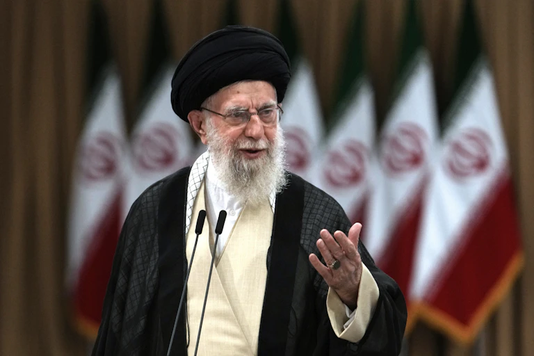 An old man in a black turban and robe with a long white beard and wearing glasses speaks in front of a microphone
