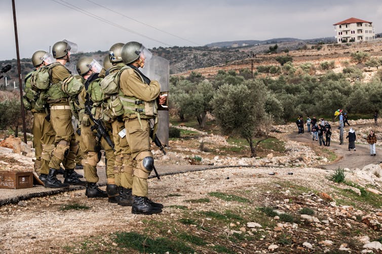 Several armed IDF soldiers on a hilltop looking down on Palestinian people.