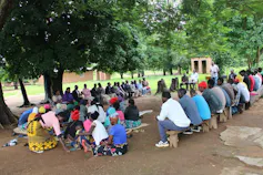 A group of about 40 people sit outside under green trees holding a discussion