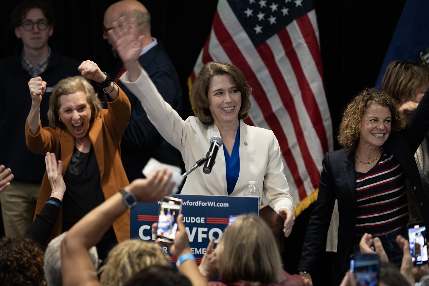 Woman in white blazer stands at podium and smiles as she waves at the crowd