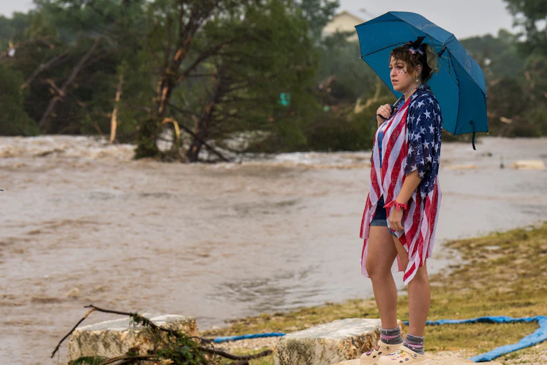 Standing on dry land, a woman holding an umbrella, wearing shorts and a U.S. flag draped around her shoulders, looks forlornly at the rushing flood waters.