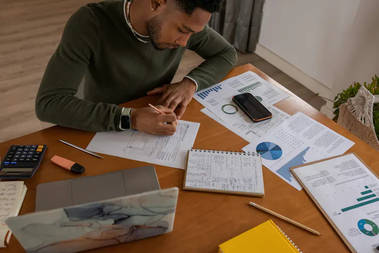A man sits at a table writing in a notebook while surrounded by graphs and charts