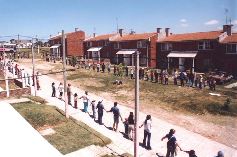 Los niños y los adultos se toman de la mano al lado de la acera frente a casas de baja altura.