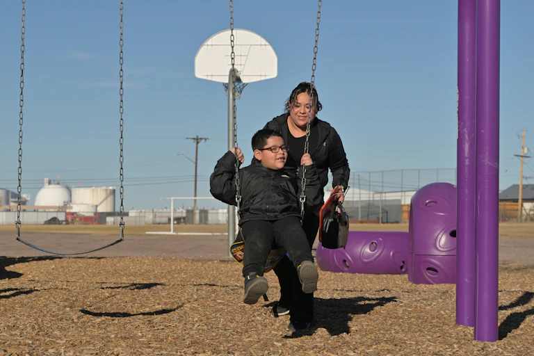 A mother pushes her young son on a swing with an oil and gas storage facility in the background.
