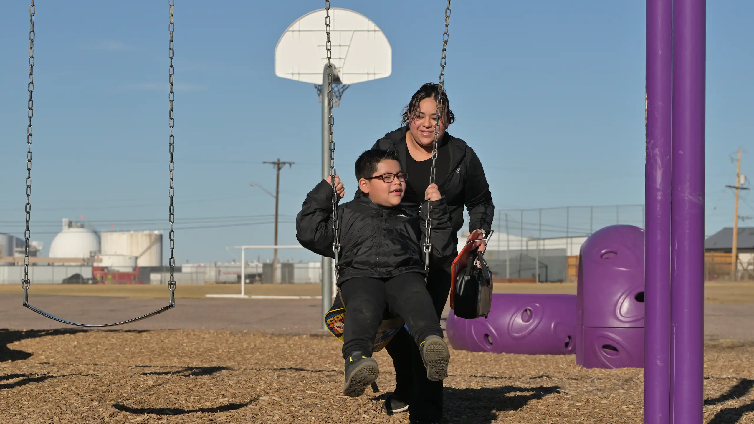 A mother pushes her young son on a swing with an oil and gas storage facility in the background.