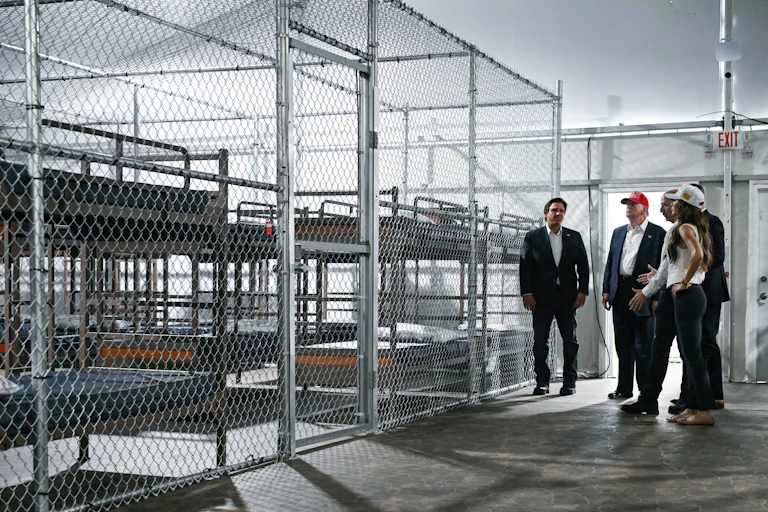 Gov. Ron Desantis, President Donald Trump and U.S. Department of Homeland Security Secretary Kristi Noem stand in front of cages full of bunk beds.