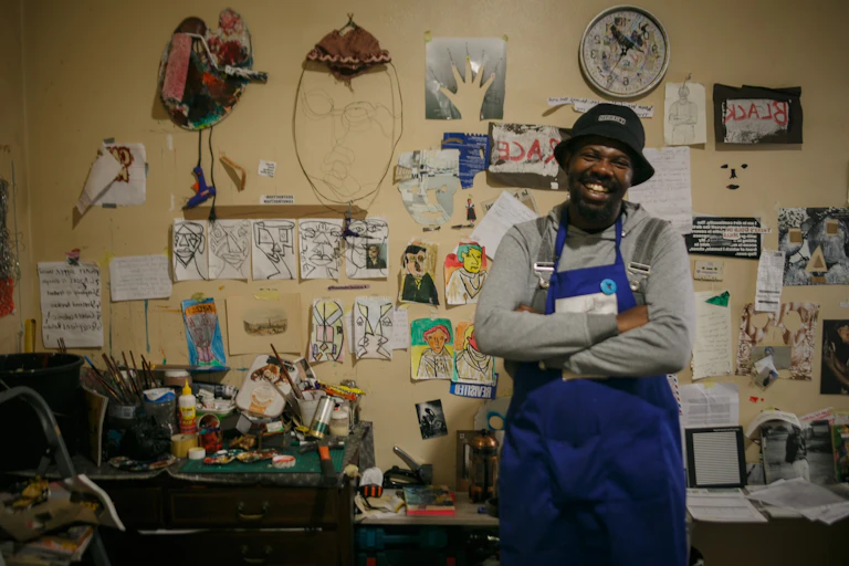 An African man in a work apron smiles broadly as he stands, arms crossed, in a studio space, walls covered in images and drawings.