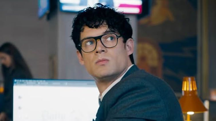 Un chico con traje, pelo rizado y gafas mira hacia arriba, fuera de campo.