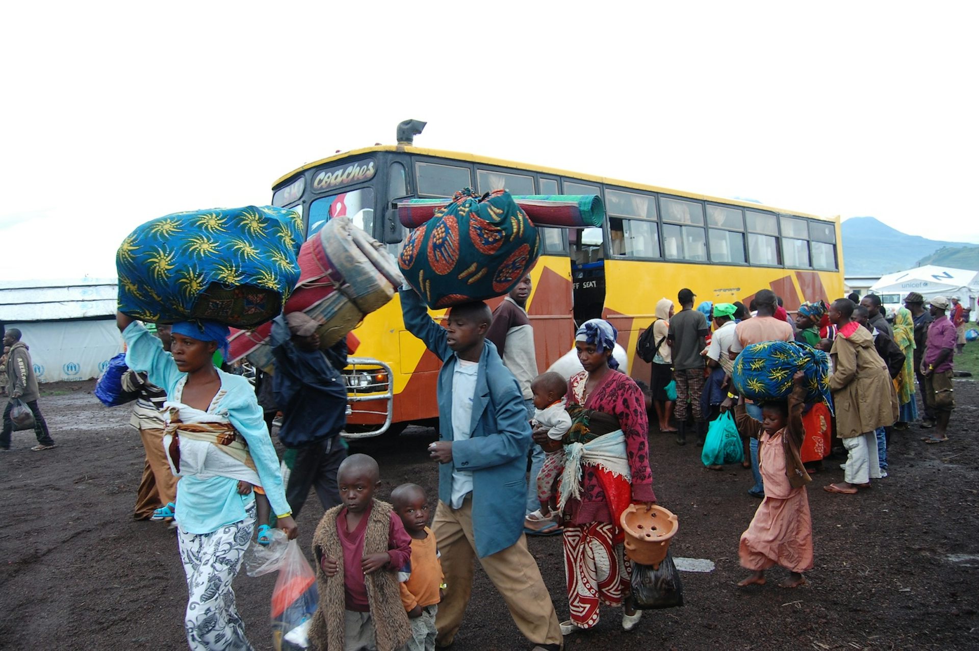 Women and children carrying bags on their heads and in their hands with a yellow bus behind them.
