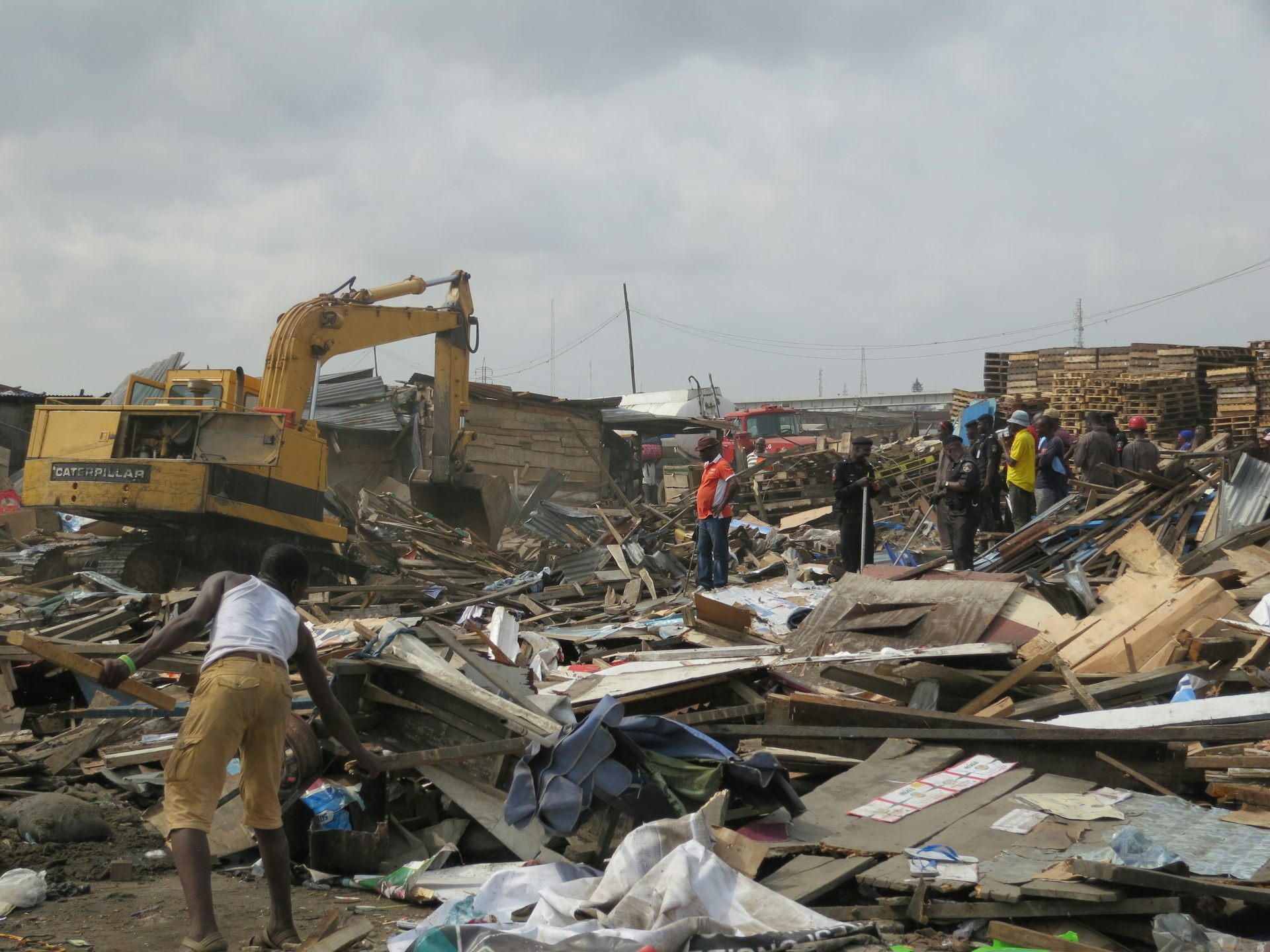 A yellow bulldozer demolishes homes, with wood and iron lying all over the ground and people trying to salvage belongings