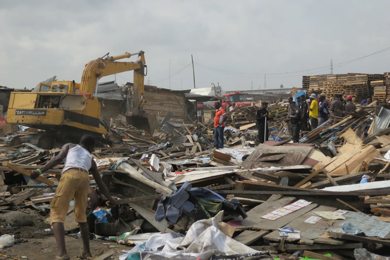 A yellow bulldozer demolishes homes, with wood and iron lying all over the ground and people trying to salvage belongings