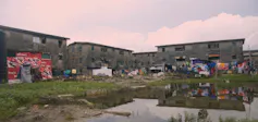 A set of four grey apartment buildings surrounded by shanties and a swampy pool of water