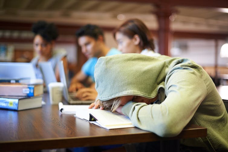 A student wearing a hooded sweatshirt slumps over a textbook.