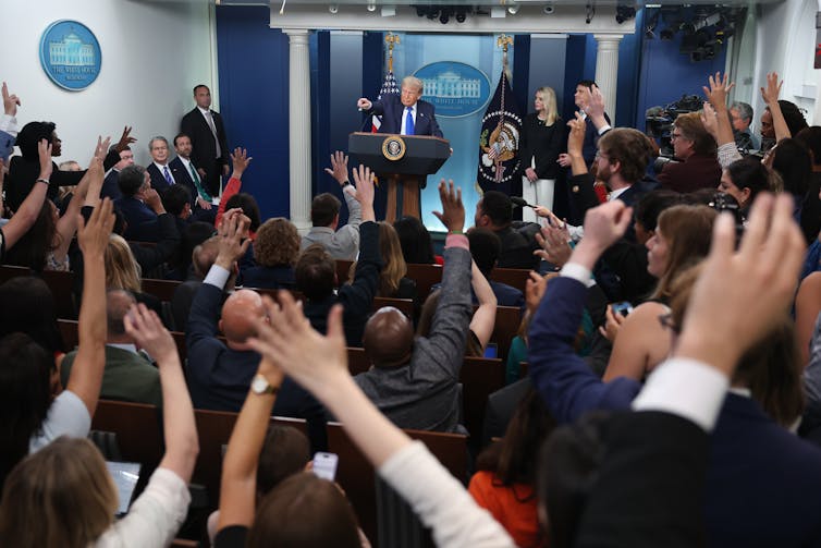 Several people raise their hands as a man at a podium answers questions.