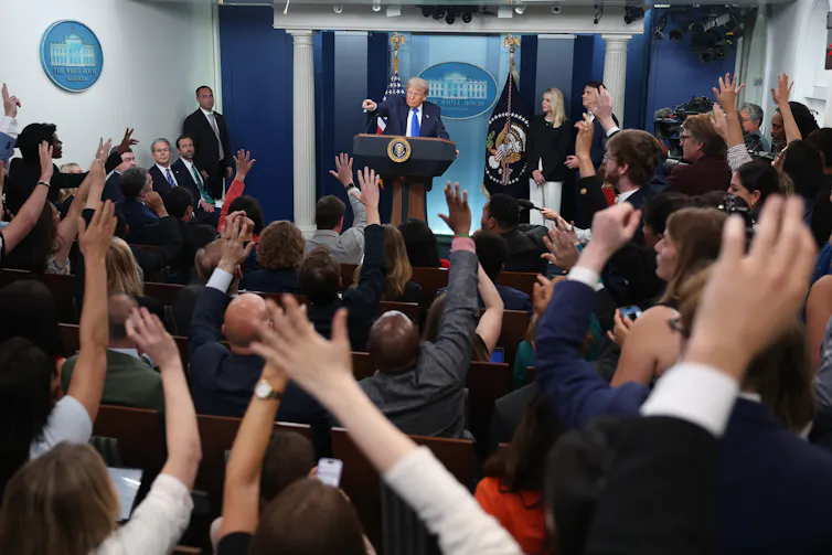 Several people raise their hands as a man at a podium answers questions.