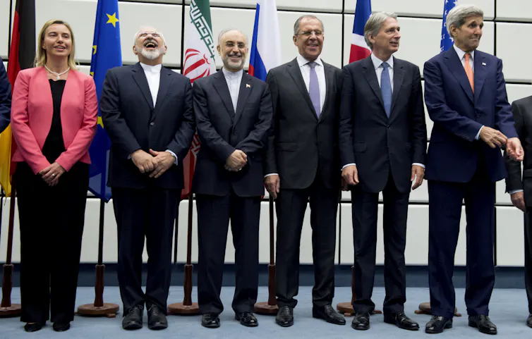 A row of men in dark suits and a woman in a pink blazer stand in a row for a photo, flags on poles are behind them