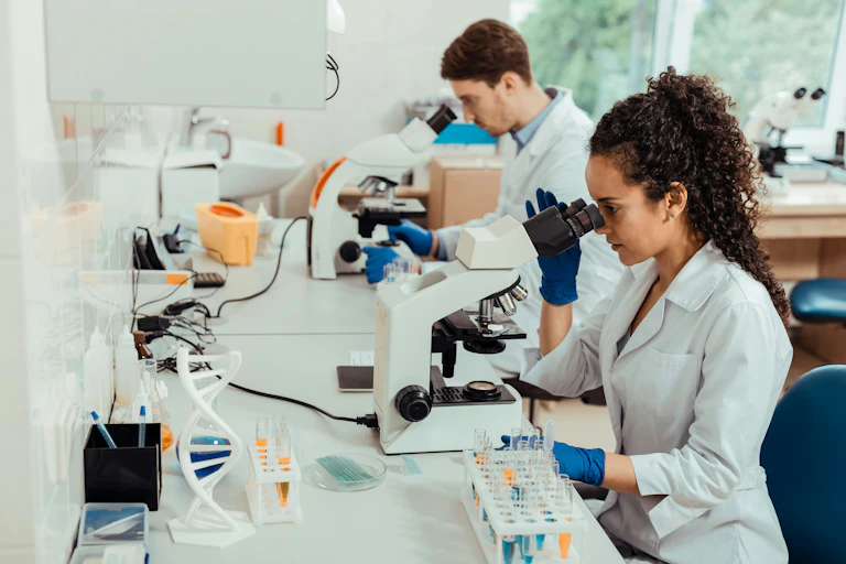 two scientists — a Black woman and a white man — in lab coats looking into microscopes