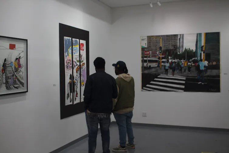 Two men with their backs to camera look at artworks displayed on walls, depicting urban themes, roads and cities.