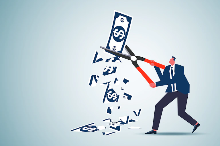A man in a suit uses a pair of scissors to snip a large bill with a dollar sign on it.