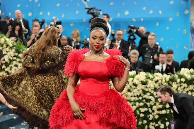 An African woman with high hair wears an opulent bright red dress that's feathered in places, a bank of news cameras in the background.