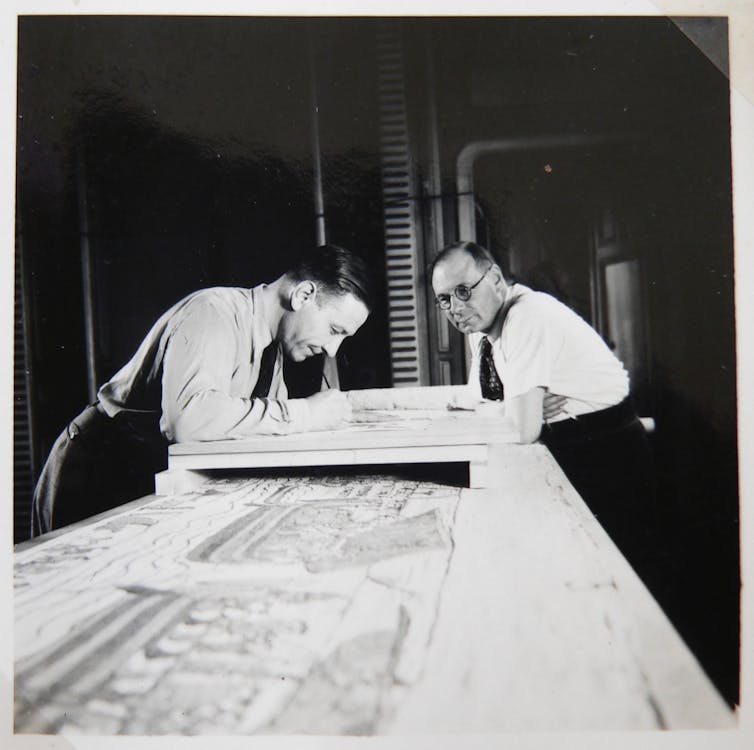 Two men looking over the Bayeux tapestry on a table.