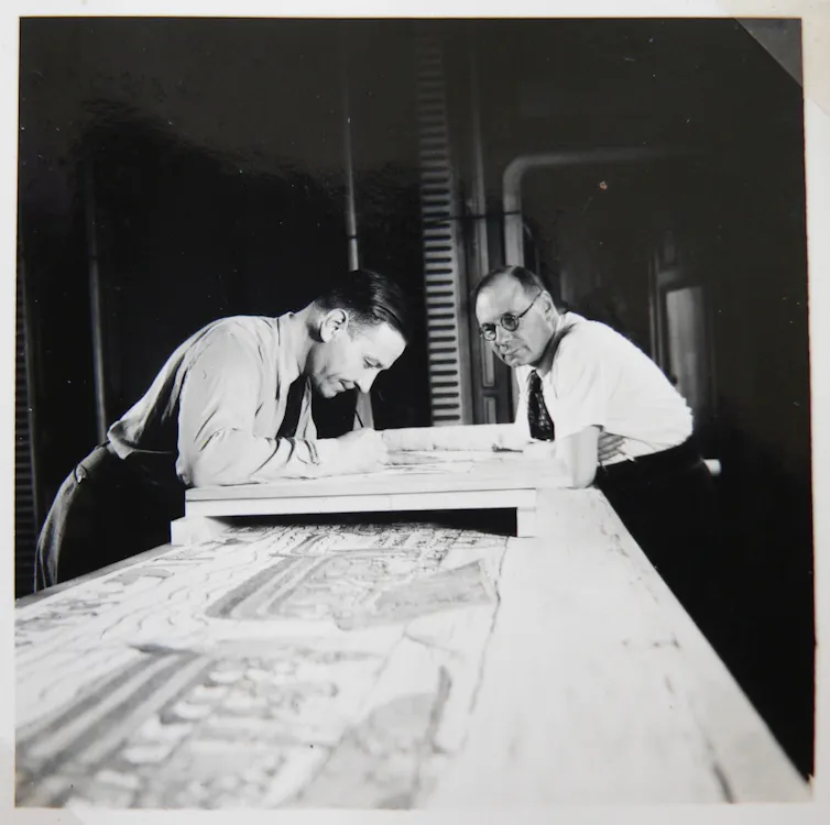 Two men looking over the Bayeux tapestry on a table.