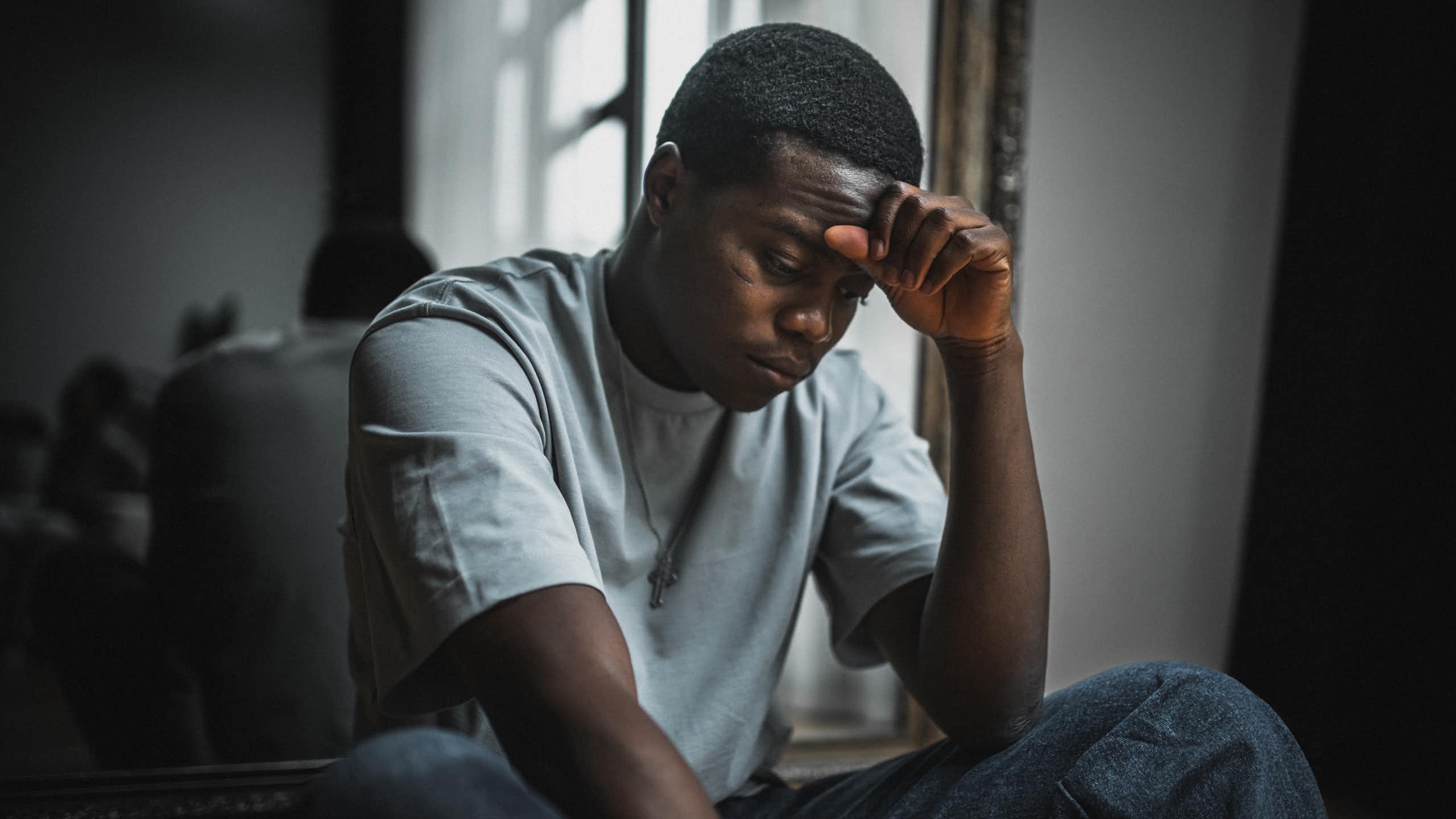 Young Black man sits while resting forehead on hand and looking stressed.