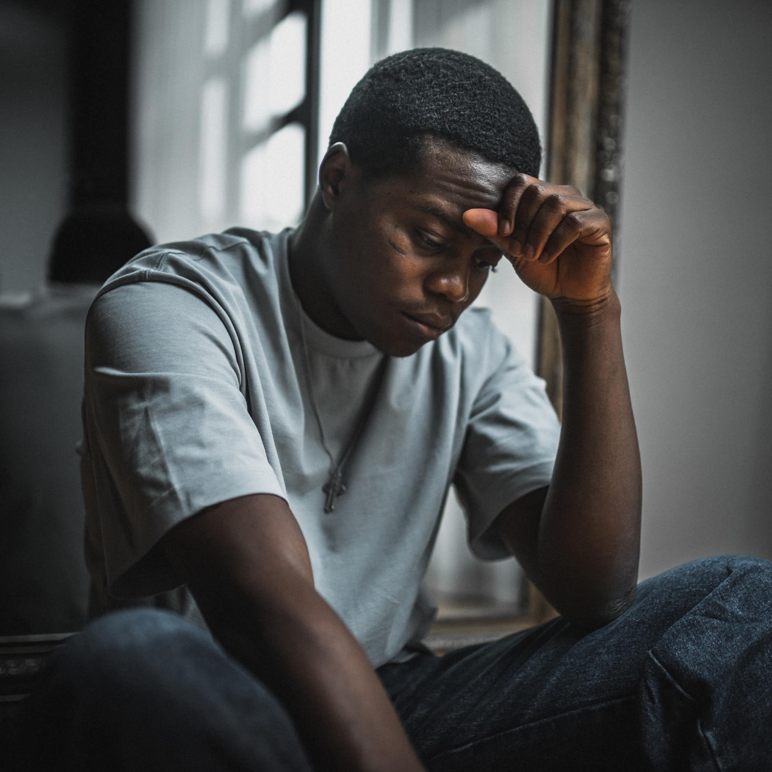 Young Black man sits while resting forehead on hand and looking stressed.