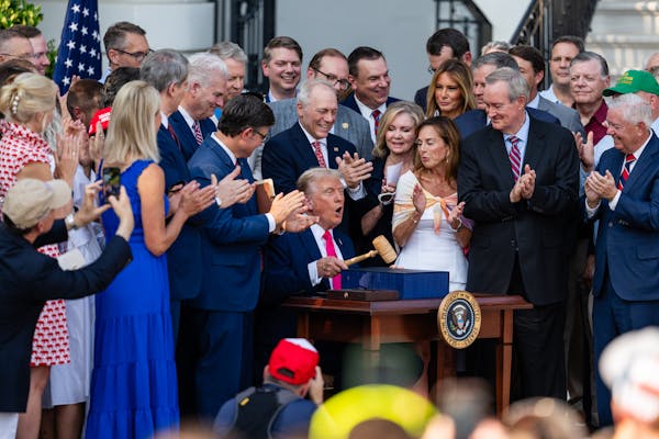 A man in suit and tie sits outdoor at a table holding a gavel as dozens of people stand behind him and clap.