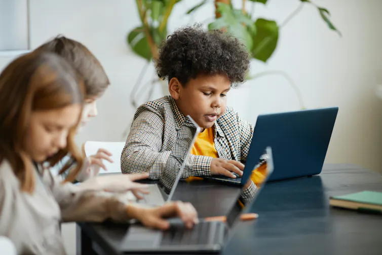 children sit at a table with laptops.