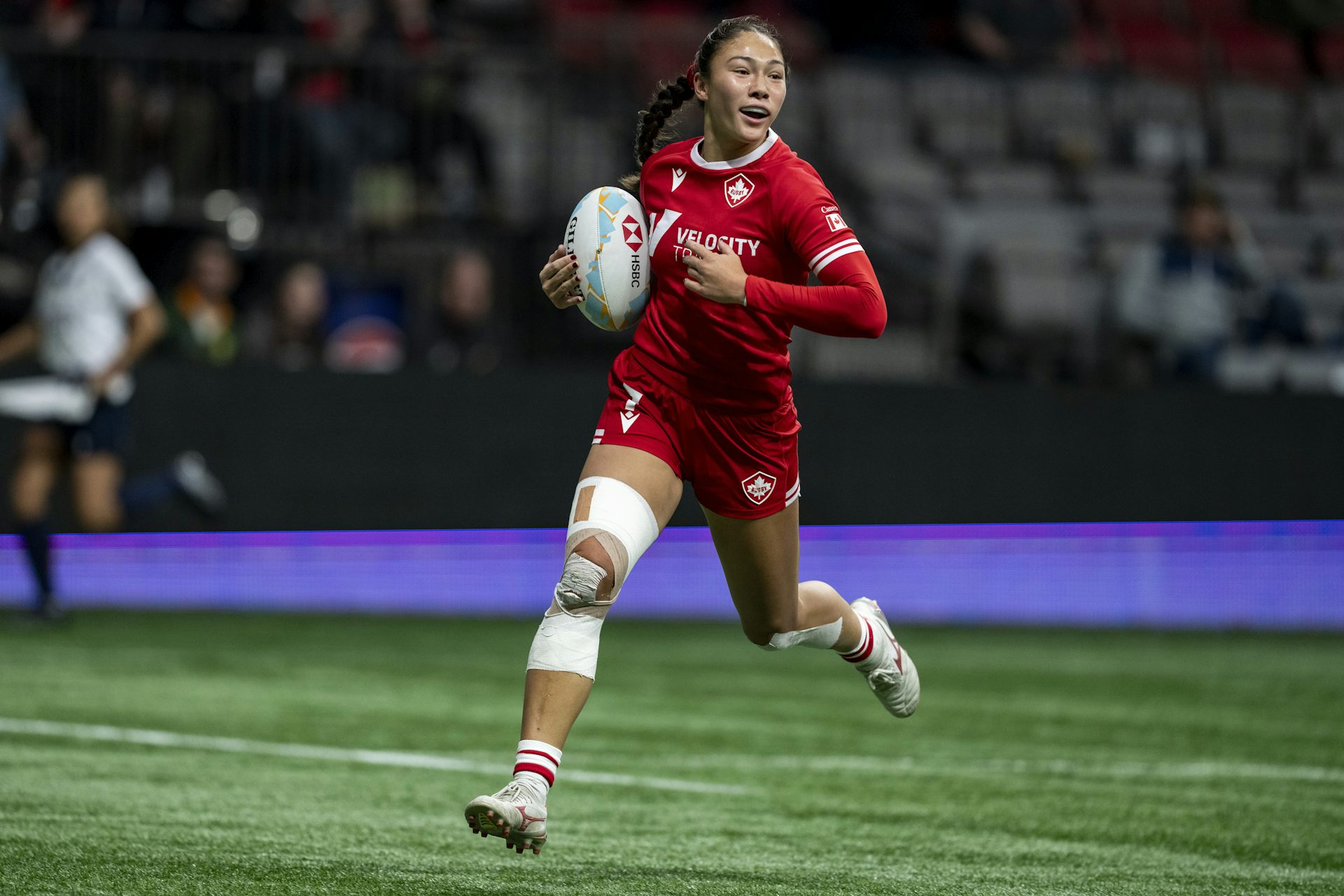 A woman in a red rugby uniform runs down a rugby pitch while holding the ball