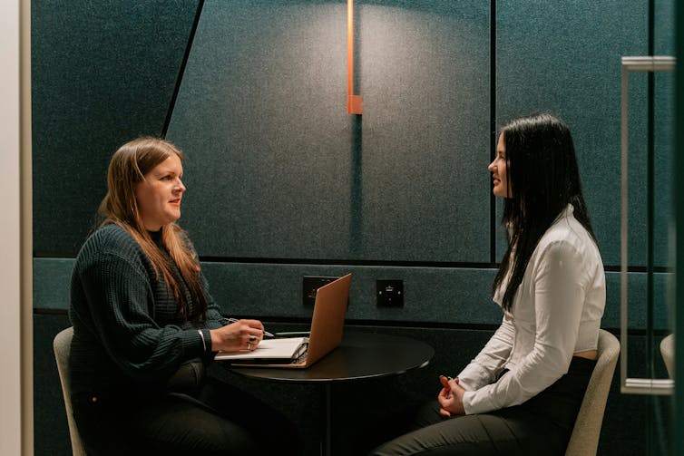 Two women have a conversation at a small table in an office setting