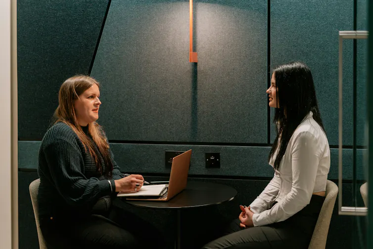 Two women have a conversation at a small table in an office setting