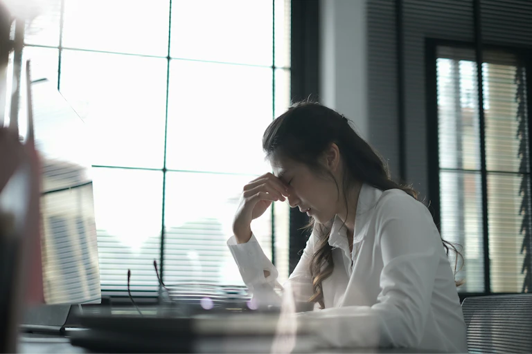 A woman sits at a desk and pinches the bridge of her nose with her eyes closed
