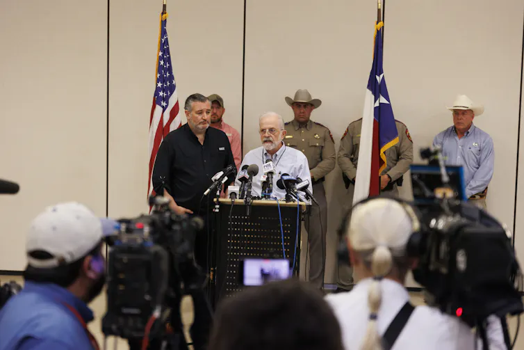 A short older man with glasses stands at podium. Three larger men in cowboy hats, two of them in uniforms, stand behind him.