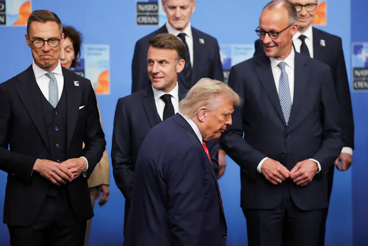 A man with blond hair walks past a group of people in suits.