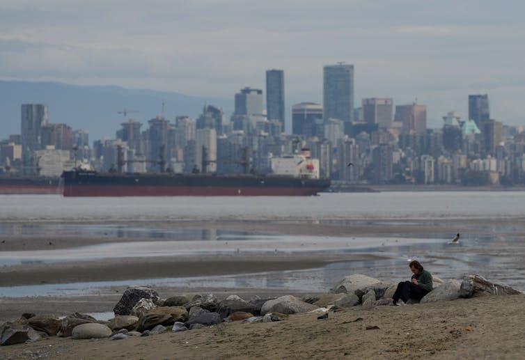 La persona está sentada en la playa de arena. El barco de carga se puede ver en el agua en la distancia, detrás de él hay un contorno de la ciudad con rascacielos