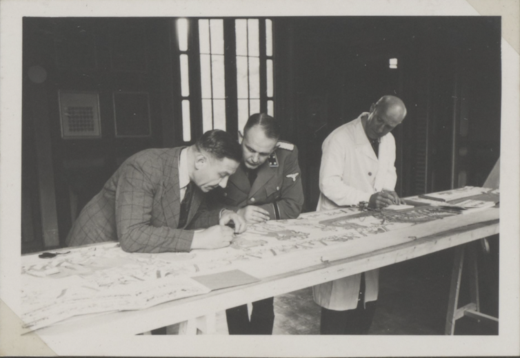 Three men stand at a long table examining something on it.