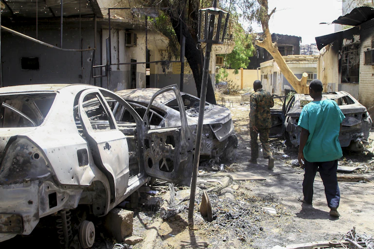 Two men walking past the burned out shells of three vehicles with a destroyed building on either side of them