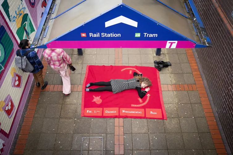 A woman lying on a pink blanket on the ground as part of a performance artwork.