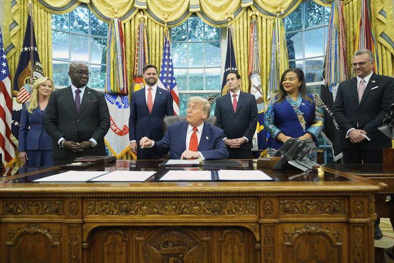 A rotund man with orange-ish white hair and facial bronzer sits at an desk in an ornate office. A woman stands to his left and a Black man to his right.