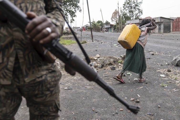 El niño usa agua al lado del soldado armado cuya arma es visible en primer plano.