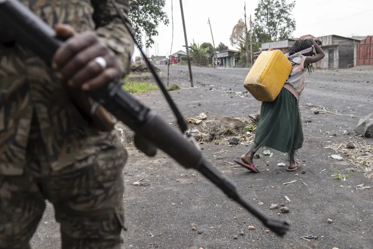 A child carries water past an armed soldier whose gun and hand is all that's visible in the foreground.